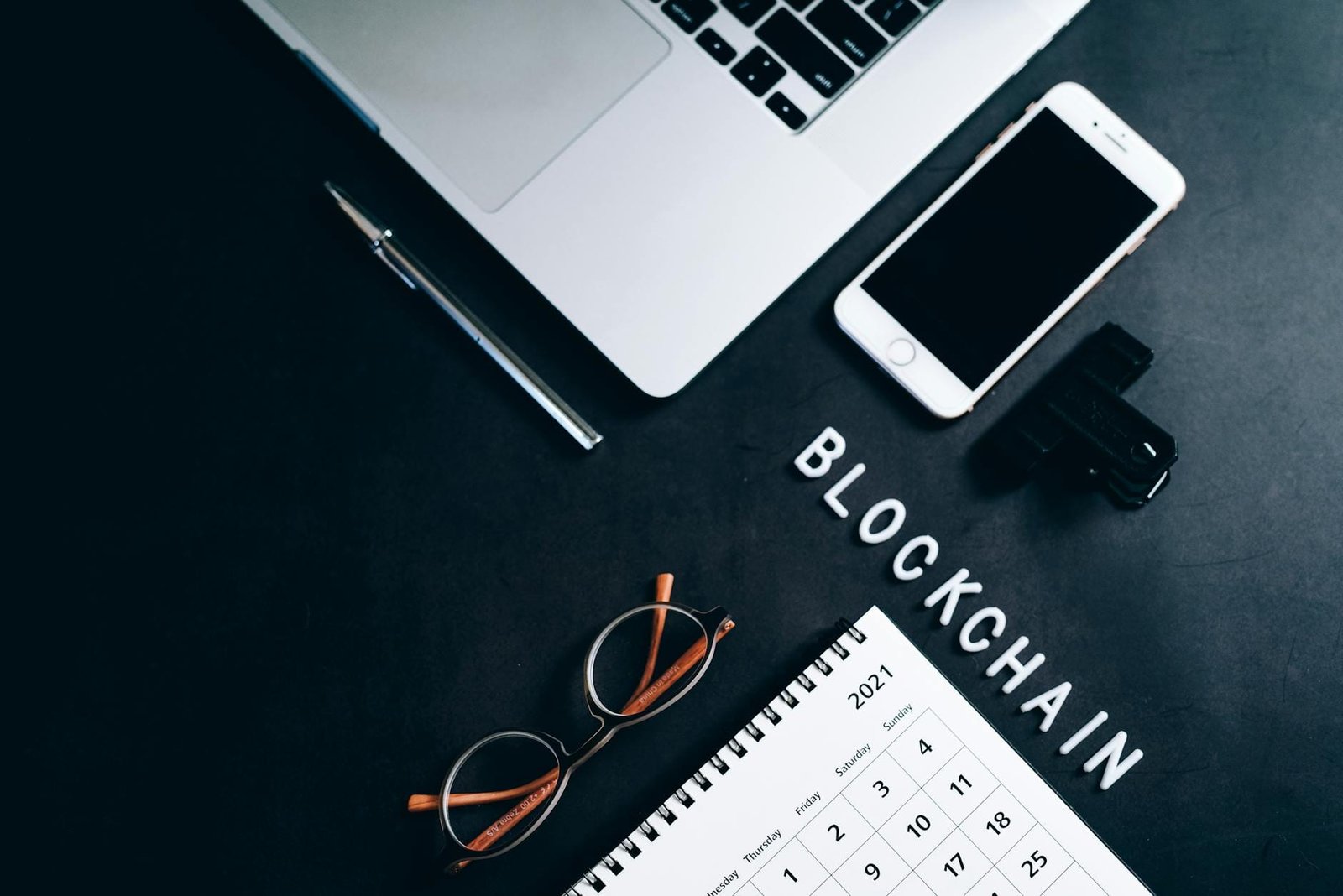 Overhead shot of a desk with a laptop, smartphone, calendar, pen, and 'BLOCKCHAIN' text.
